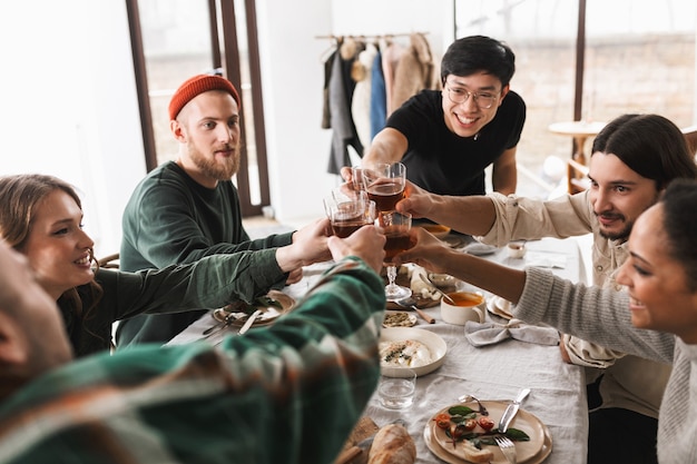 Foto il giovane uomo asiatico allegro in occhiali e maglietta nera applaude felicemente i vetri di vino con i colleghi