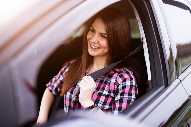 Young joyful beautiful girl is buckling up in a drivers seat of her car.