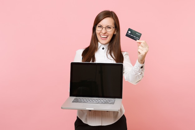 Giovane donna d'affari gioiosa con gli occhiali in possesso di carta di credito, computer pc portatile con schermo vuoto vuoto isolato su sfondo rosa. signora capo. ricchezza di carriera di successo. copia spazio per la pubblicità.