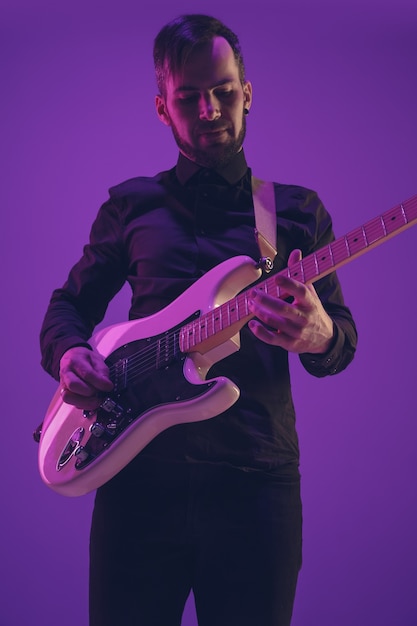 Giovane e gioioso musicista caucasico che suona la chitarra su sfondo viola sfumato per studio alla luce al neon. concetto di musica, hobby, festival. ritratto colorato di artista moderno. attento e ispirato.