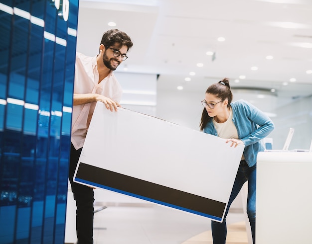 Young joyful couple is carrying a brand new unpacked TV bought in a very bright tech store.