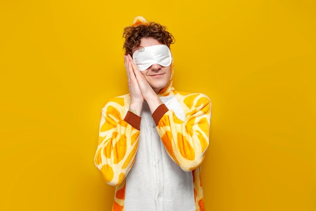 Young joyful guy in funny baby giraffe pajamas and sleep mask is sleeping on yellow background