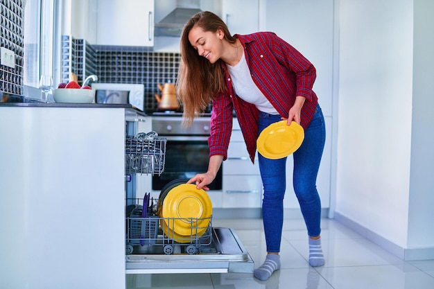 Young joyful happy satisfied smiling cute housewife woman using modern dishwasher for wash dishes and glasses at white home kitchen