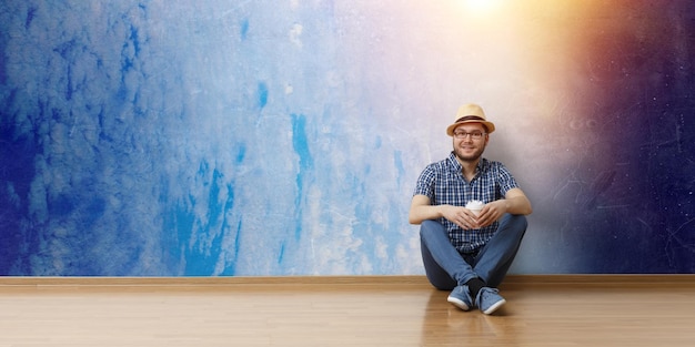 Young joyful man in casual clothes and hat sitting on floor, space background. mixed media