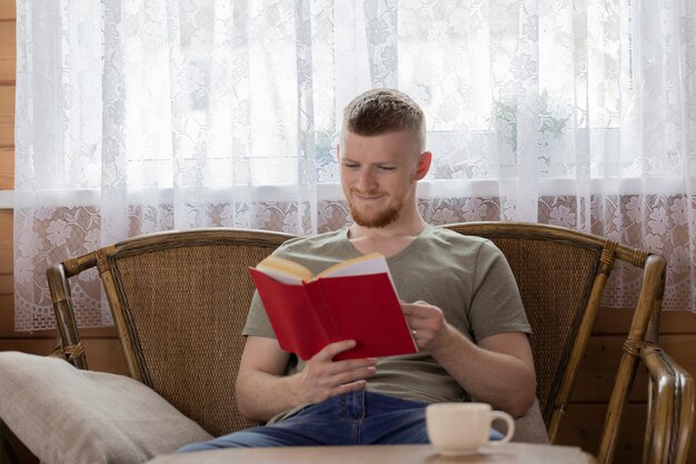 Молодой радостный человек читает книгу на плетеной скамейке в загородном доме