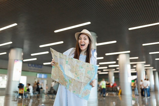 Young joyful traveler tourist woman in hat holding paper map, searching route while waiting in lobby hall at international airport