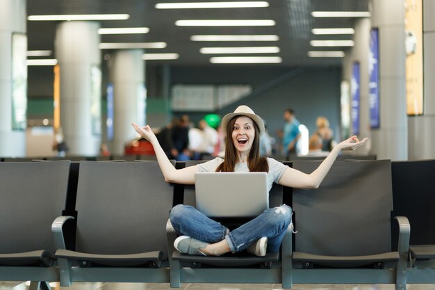 Young joyful traveler tourist woman with laptop sitting with crossed legs, meditate, spread hands, waiting in lobby hall at airport