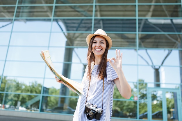 Young joyful traveler tourist woman with retro vintage photo camera holds paper map, show OK gesture at international airport