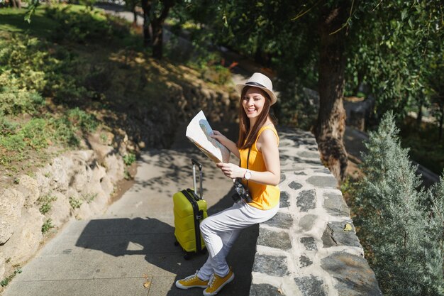노란색 옷을 입은 젊은 여행자 관광 여성, 여행 가방이 달린 모자는 도시 야외의 돌 위에 앉아 있는 도시 지도를 들고 있습니다. 주말 휴가를 여행하기 위해 해외로 여행하는 소녀. 관광 여행 라이프 스타일.