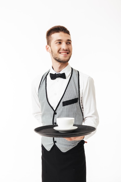 Фото Молодой радостный официант в униформе, счастливо держащий поднос с чашкой кофе в руке