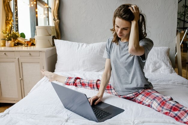 Young joyful woman in pajama sitting on bed using on laptop at home