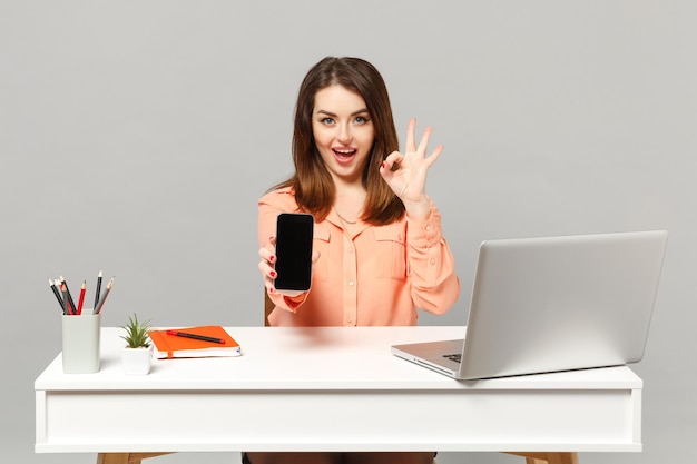 Молодая радостная женщина показывает жест ОК, держа мобильный телефон с пустым пустым экраном, сидит за столом с ноутбуком, изолированным на сером фоне. Достижение концепции деловой карьеры. Скопируйте пространство для копирования.