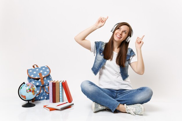 Young joyful woman student with headphones listening music pointing index fingers up sitting near globe backpack school books isolated