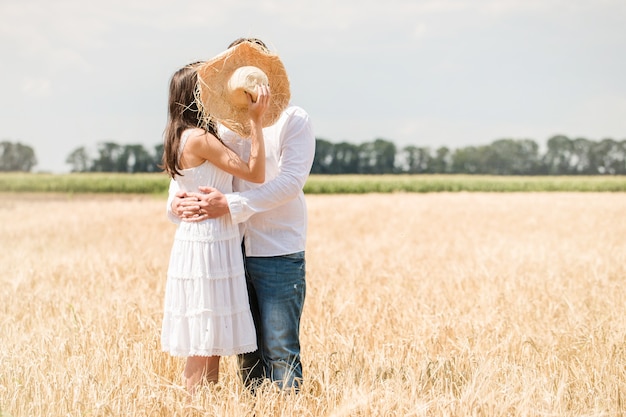 Young kissing couple hiding behind hat in the field