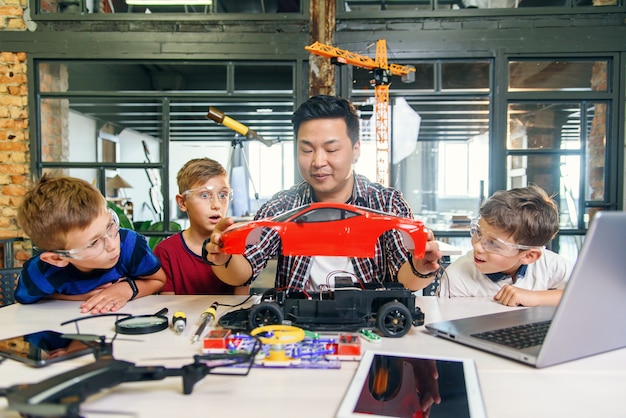 Young korean man of electronics engineer with young children\
using screwdriver to disassemble robotic machine at the table in\
the modern school. slow motion