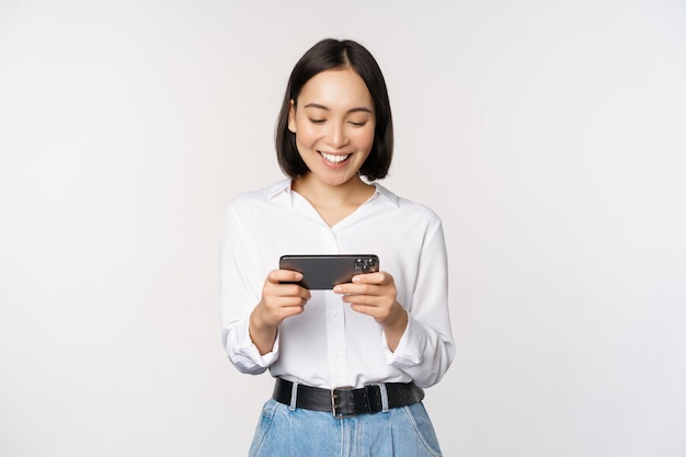흰색 배경 위에 서 있는 가로 전화 화면을 보고 스마트폰으로 모바일 비디오 게임을 하는 젊은 한국 여성 아시아 소녀