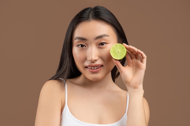 Young korean woman with clean skin holding lime half near her face posing with citrus fruit over