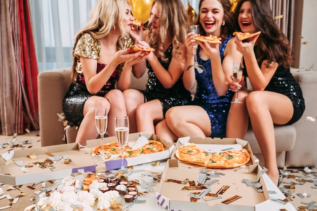 Giovani donne che celebrano la festa di compleanno a casa, mangiano pizza, bevono champagne, si divertono. coriandoli intorno.