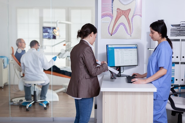 Young lady asking informations filling in stomatological form while patients talking sitting on chair in waiting area