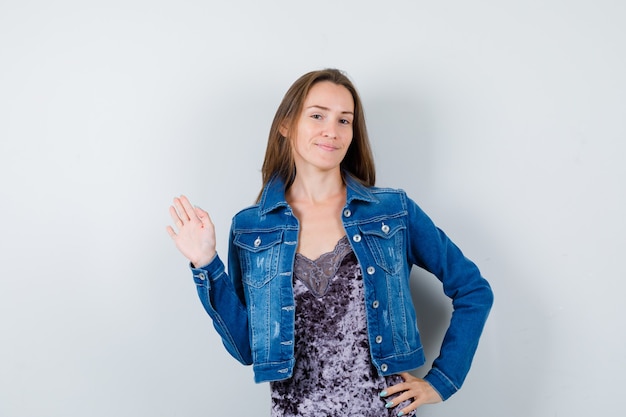 Young lady in blouse, denim jacket waving hand for greeting while keeping hand on hip and looking happy , front view.