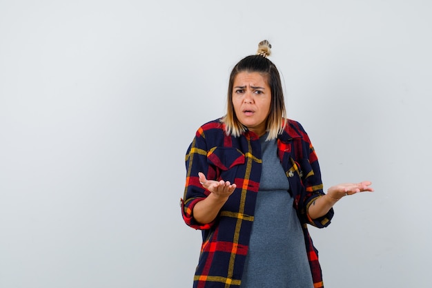 Young lady in casual checked shirt making asking question gesture and looking wistful , front view.