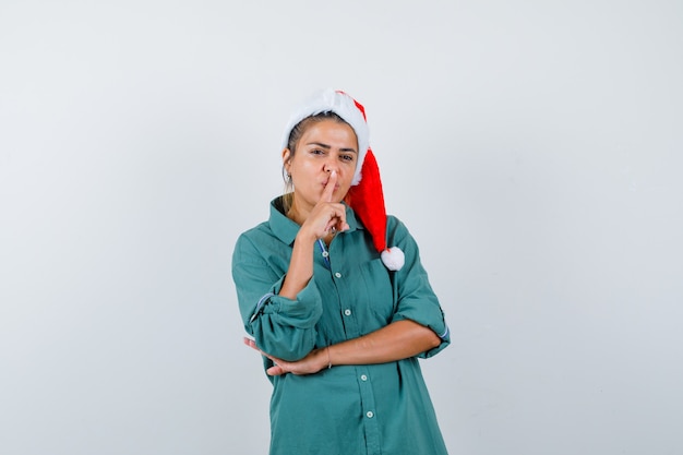 Young lady in christmas hat, shirt showing silence gesture and looking careful , front view.