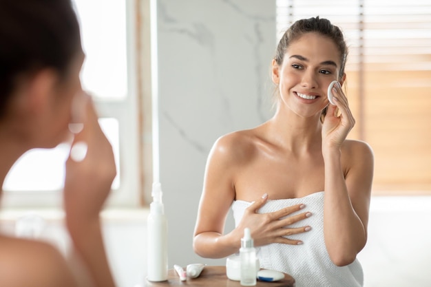 Giovane donna che pulisce il viso utilizzando un batuffolo di cotone in piedi in bagno