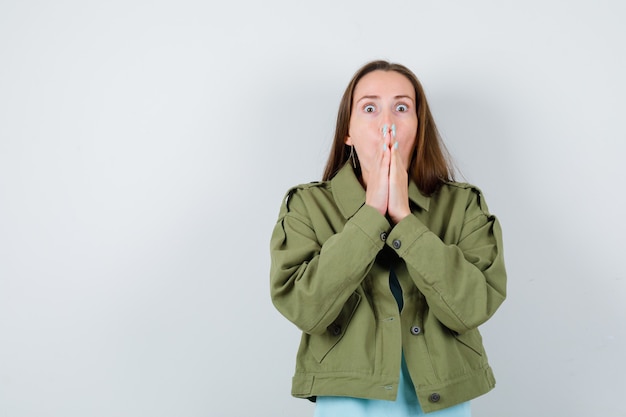 Young lady covering mouth with hands in t-shirt, jacket and looking scared , front view.