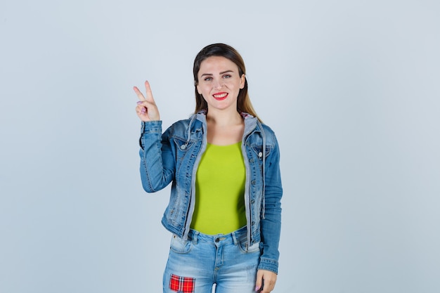Young lady in denim outfit showing victory gesture and looking joyful , front view.