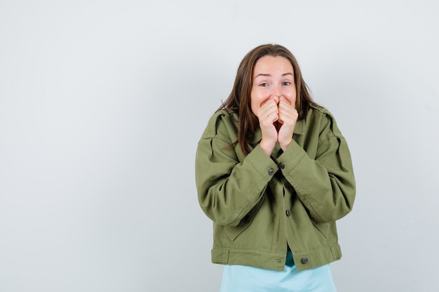 Young lady in green jacket with hands on mouth and looking glad , front view.