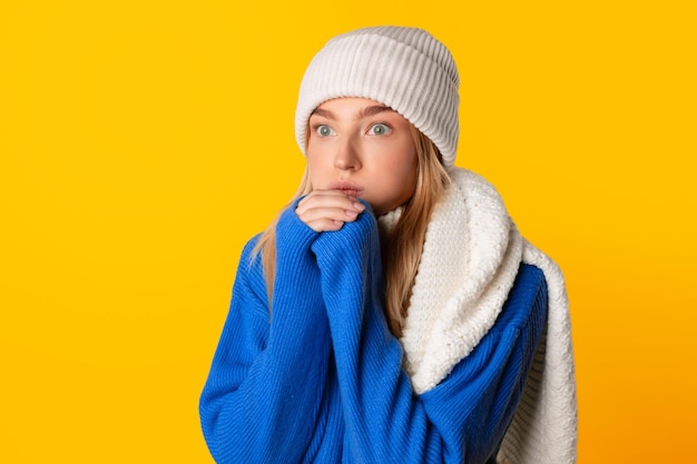 Young lady is frozen and warming hands in winter attire yellow background