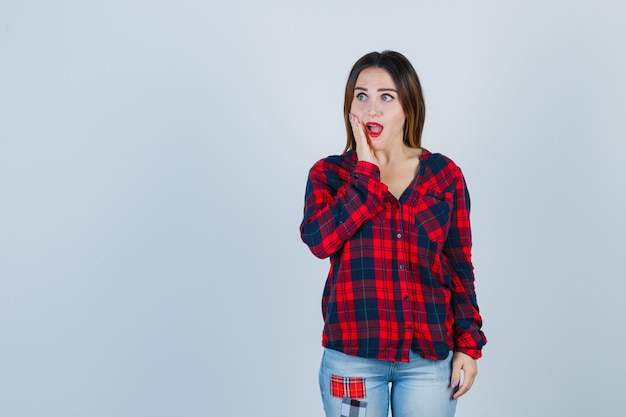 Young lady keeping hand on cheek, opening mouth in checked shirt, jeans and looking astonished , front view.