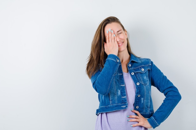 Young lady keeping hand on eye in t-shirt, jacket and looking merry. front view.