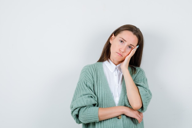 Young lady leaning cheek on palm in shirt, cardigan and looking pensive. front view.