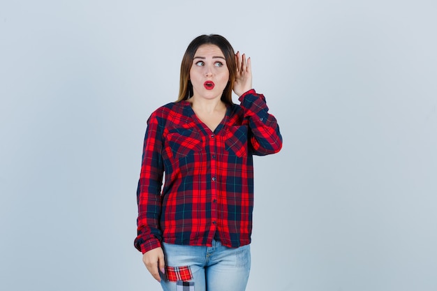 Young lady overhearing private conversation by keeping hand behind ear in checked shirt, jeans and looking shocked. front view.