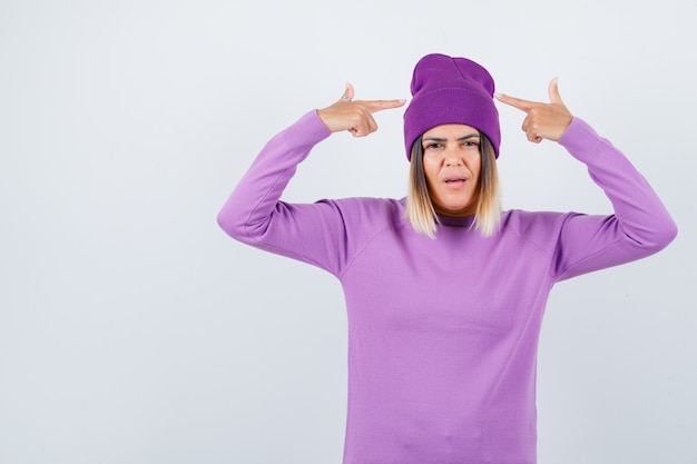 Giovane donna che indica il suo berretto in maglione viola, berretto e sembra fiducioso, vista frontale.