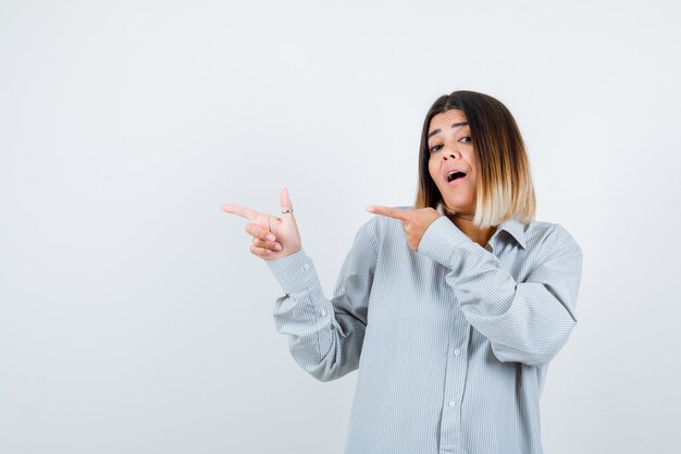 Young lady pointing to the left side in oversized shirt and looking puzzled. front view.