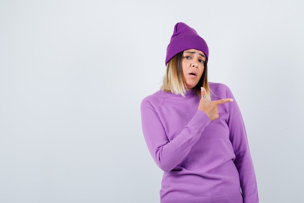 Photo young lady pointing to the right side in purple sweater, beanie and looking anxious. front view.