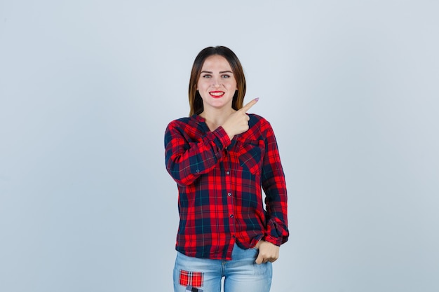 Giovane donna che indica nell'angolo in alto a destra in camicia a quadri, jeans e sembra allegra. vista frontale.