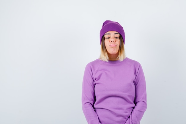 Молодая дама позирует, дует губы в фиолетовом свитере, шапочке и выглядит удивленным, вид спереди.