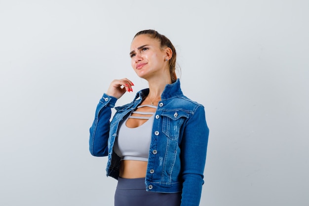 Young lady posing while looking away, raising hand in top, denim jacket and looking focused , front view.