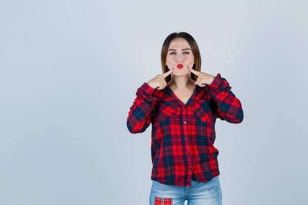 Young lady pouting lips, keeping fingers on cheeks in checked shirt, jeans and looking joyful. front view.