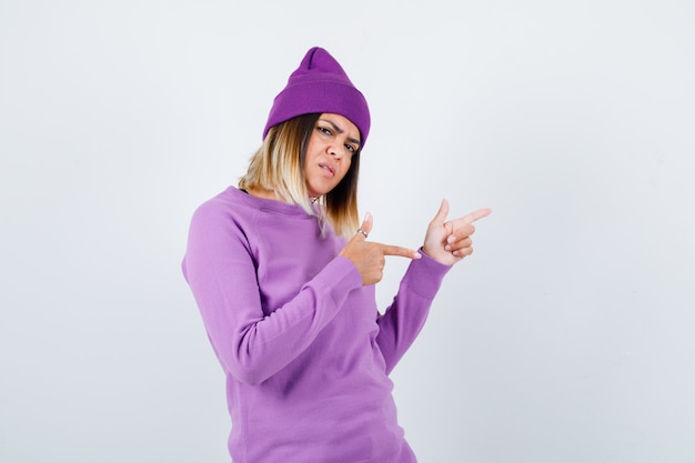 Young lady in purple sweater, beanie pointing to the right side and looking serious , front view.