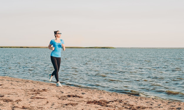 実行している若い女性。日当たりの良い夏の砂のビーチで実行されている女性ランナー。海海海岸近くのトレーニング。美しいフィットの女の子。フィットネスモデル白人民族屋外。減量運動。ジョギング。