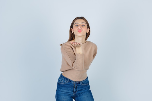 Young lady sending air kiss in beige sweater, jeans and looking pretty. front view.