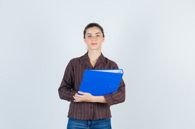 Young lady in shirt, jeans holding folder, looking at camera and looking serious , front view.