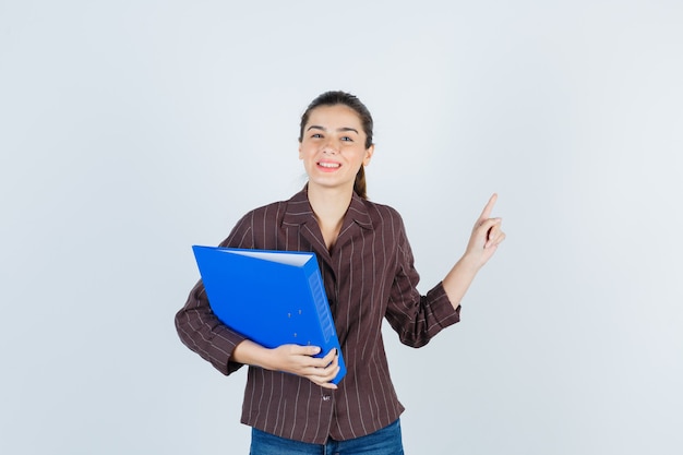 Young lady in shirt, jeans holding folder, pointing up and looking happy , front view