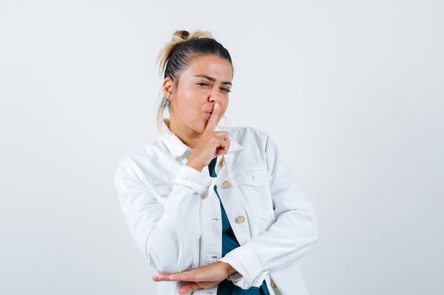 Young lady in shirt, white jacket showing silence gesture and looking careful , front view.