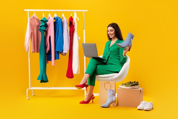 Молодая женщина-продавец, держащая элегантную обувь и пользующаяся ноутбуком, сидит возле вешалки для одежды и получает онлайн-заказы