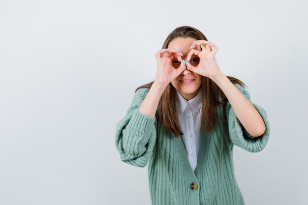 Young lady showing binoculars gesture in shirt, cardigan and looking funny. front view.